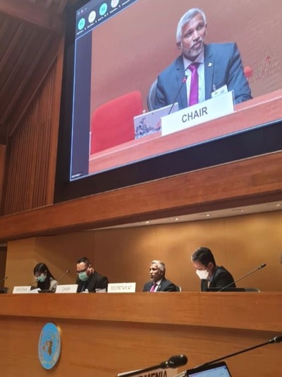 Mohamed Shareef at the chair's place on the dais of a UN conference room, shown on the large screen above behind a nameplate reading Chair