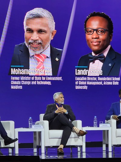 Mohamed Shareef seated in an armchair on a forum stage, beneath a large screen introducing him as former Minister of State for Environment, Climate Change and Technology, Maldives